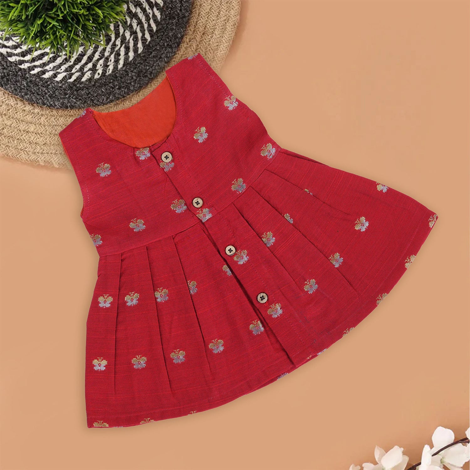 Red silk sleeveless ethnic frock for baby girls with floral woven motifs and wooden button closure, laid flat on a beige background with white florals and rustic decor.