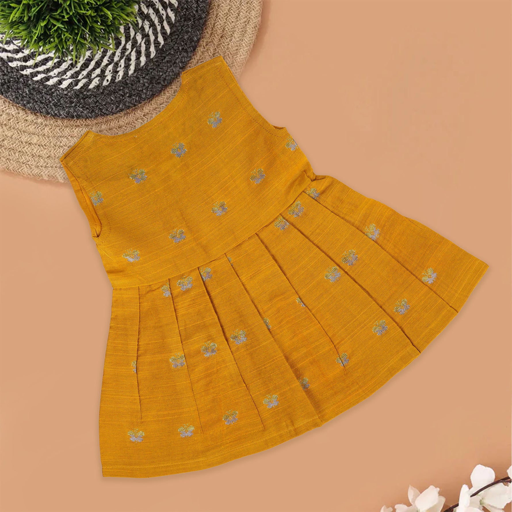 Yellow silk sleeveless ethnic frock for baby girls with floral woven motifs and wooden button closure, displayed on a peach background with floral and natural props.