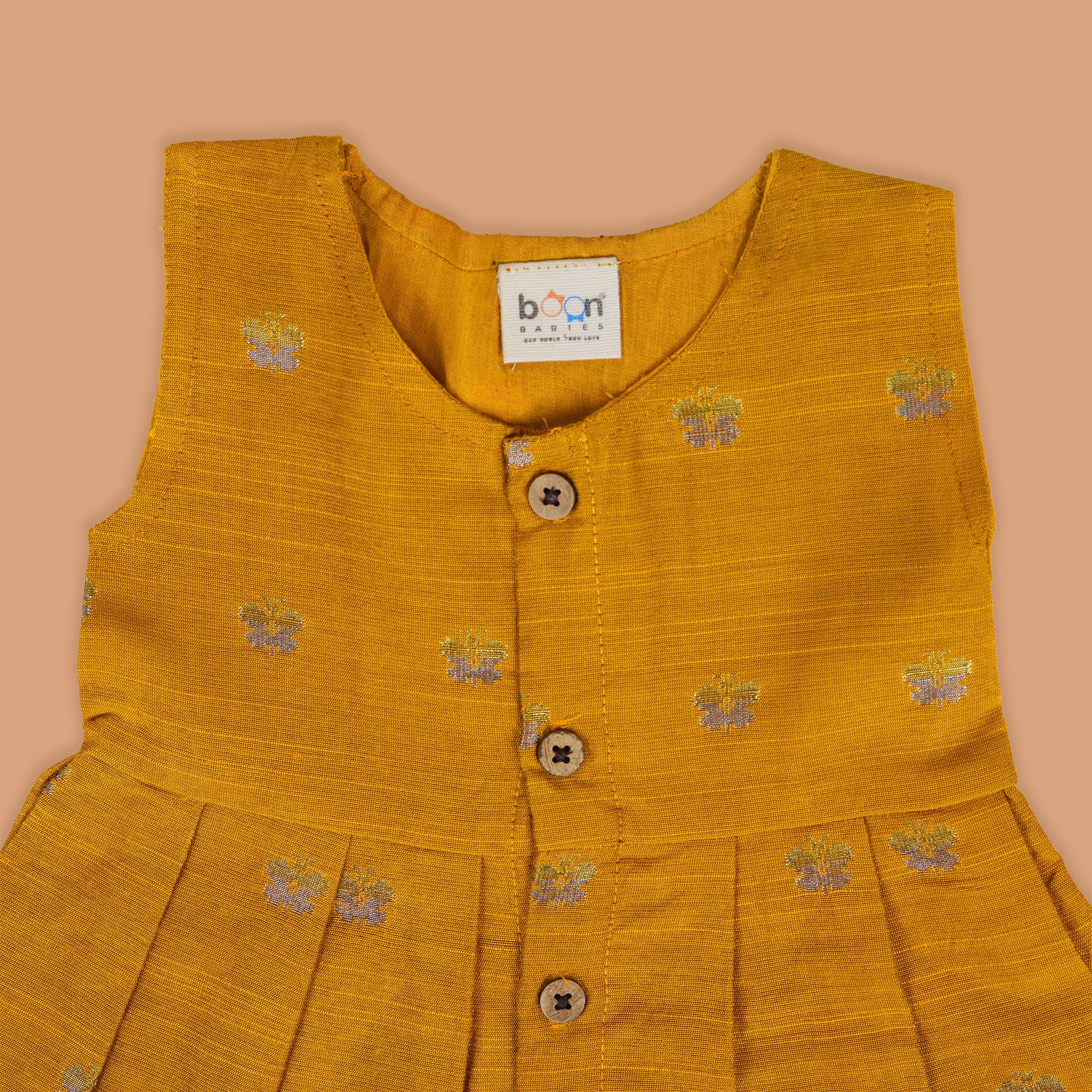Yellow silk sleeveless ethnic frock for baby girls with floral woven motifs and wooden button closure, displayed on a peach background with floral and natural props.
