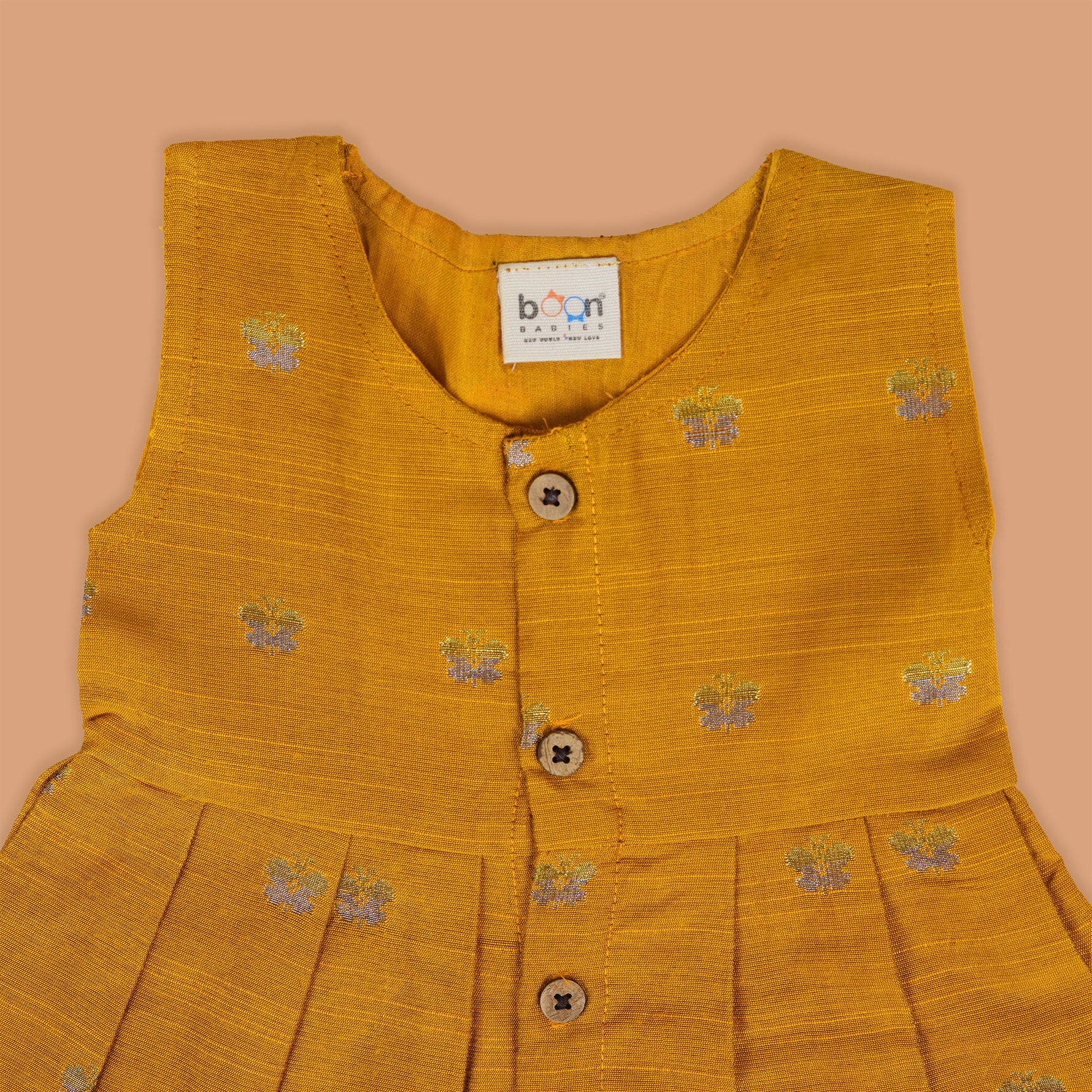 Yellow silk sleeveless ethnic frock for baby girls with floral woven motifs and wooden button closure, displayed on a peach background with floral and natural props.