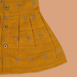 Yellow silk sleeveless ethnic frock for baby girls with floral woven motifs and wooden button closure, displayed on a peach background with floral and natural props.