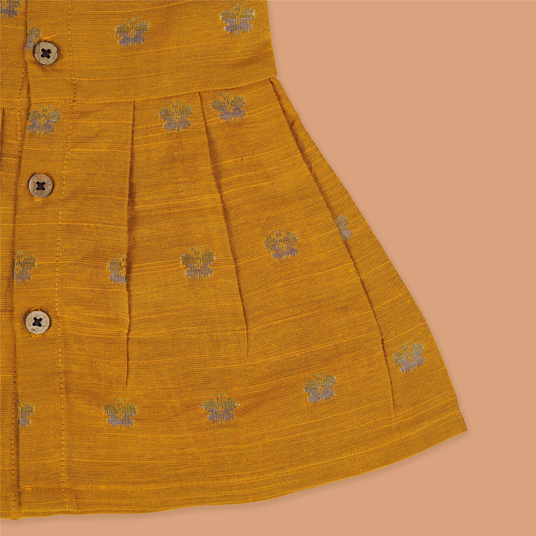 Yellow silk sleeveless ethnic frock for baby girls with floral woven motifs and wooden button closure, displayed on a peach background with floral and natural props.