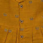 Yellow silk sleeveless ethnic frock for baby girls with floral woven motifs and wooden button closure, displayed on a peach background with floral and natural props.