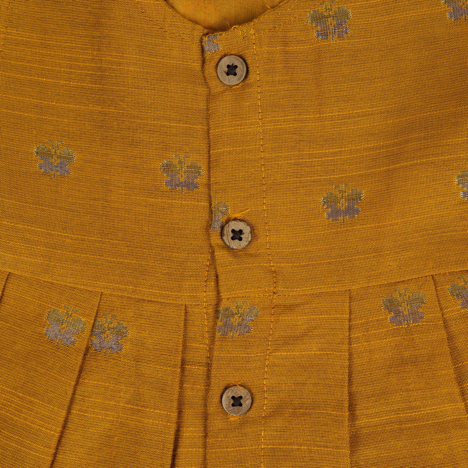 Yellow silk sleeveless ethnic frock for baby girls with floral woven motifs and wooden button closure, displayed on a peach background with floral and natural props.