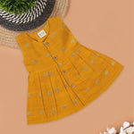 Yellow silk sleeveless ethnic frock for baby girls with floral woven motifs and wooden button closure, displayed on a peach background with floral and natural props.