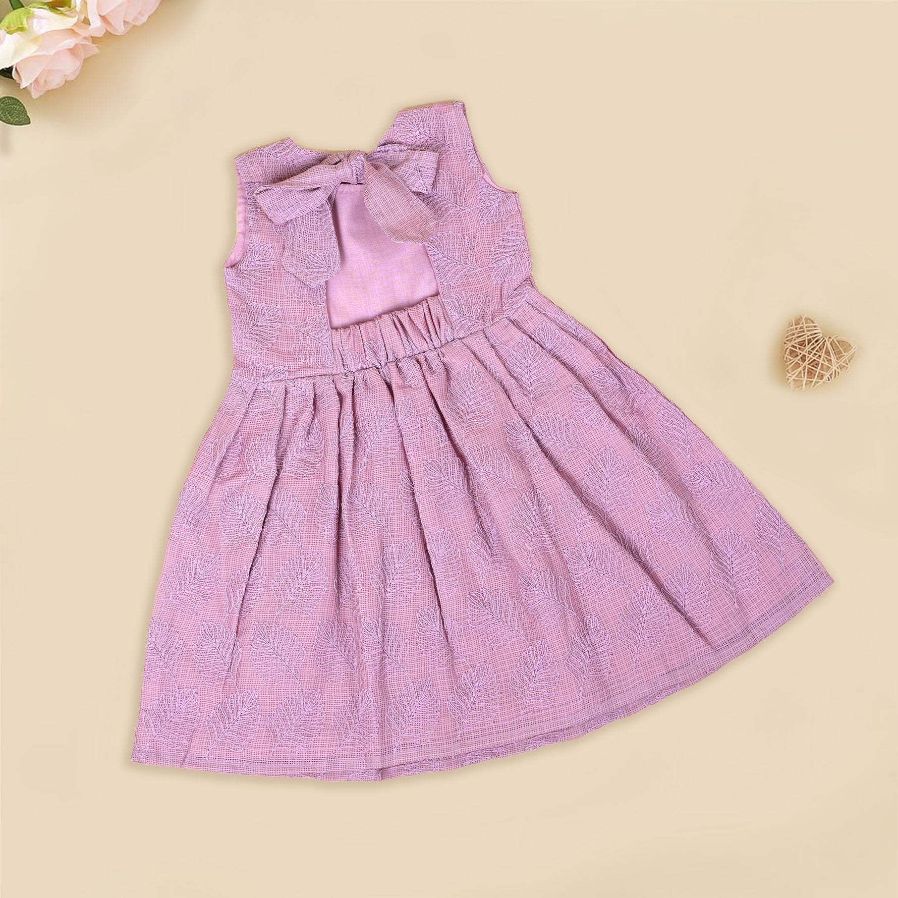 Mauve Hakoba frock for baby girls, sleeveless and pleated with soft embroidery, laid flat on a beige background beside floral accents and a heart-shaped wicker prop.