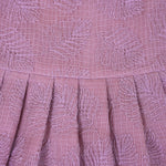 Mauve Hakoba frock for baby girls, sleeveless and pleated with soft embroidery, laid flat on a beige background beside floral accents and a heart-shaped wicker prop.