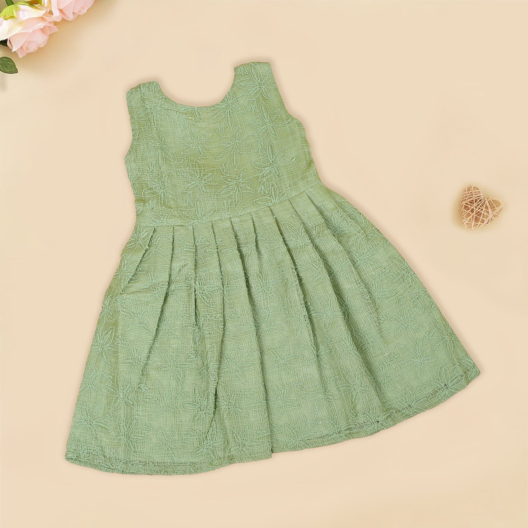 Pastel green sleeveless Hakoba frock for baby girls, featuring floral embroidery and pleated skirt design. Lightweight cotton fabric, displayed flat against a soft beige background with a decorative heart and flowers.