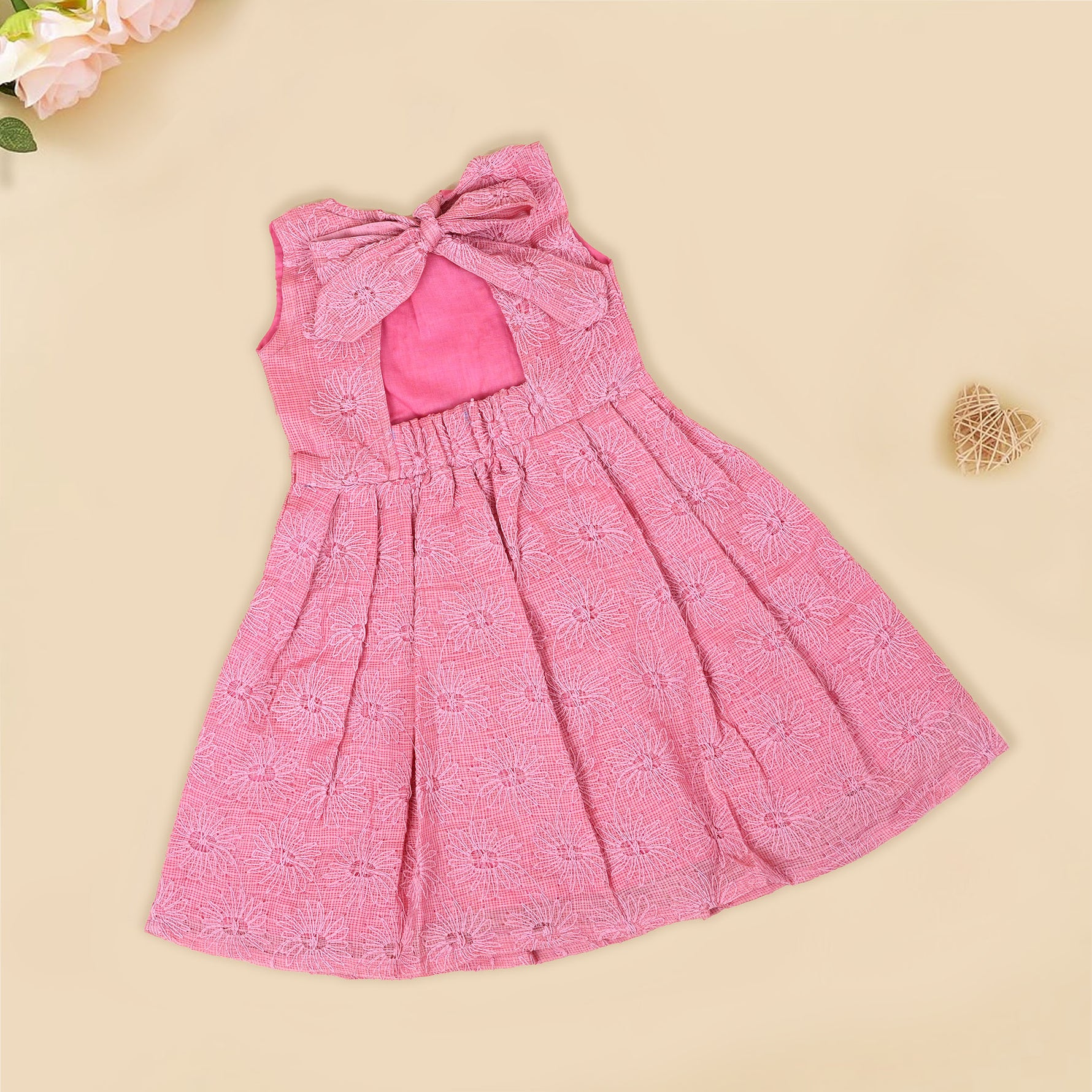 Pink sleeveless Hakoba frock for baby girls, featuring floral embroidery and pleated skirt design, laid flat on a beige background with soft floral props.