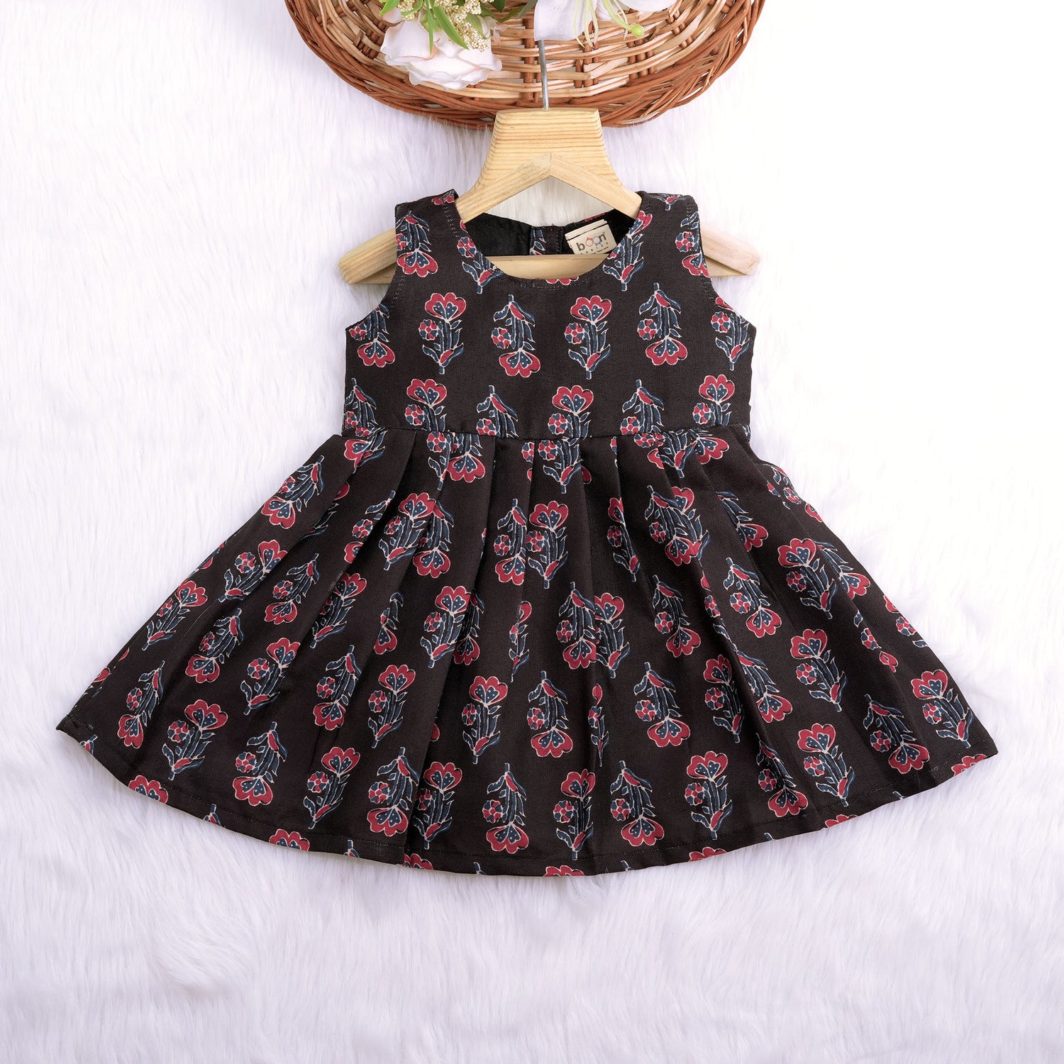 Black sleeveless slub cotton frock for baby girls with red floral block prints and a pleated flare, displayed flat on a white fur background with floral basket hanger.