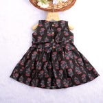 Black sleeveless slub cotton frock for baby girls with red floral block prints and a pleated flare, displayed flat on a white fur background with floral basket hanger.