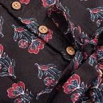 Black sleeveless slub cotton frock for baby girls with red floral block prints and a pleated flare, displayed flat on a white fur background with floral basket hanger.