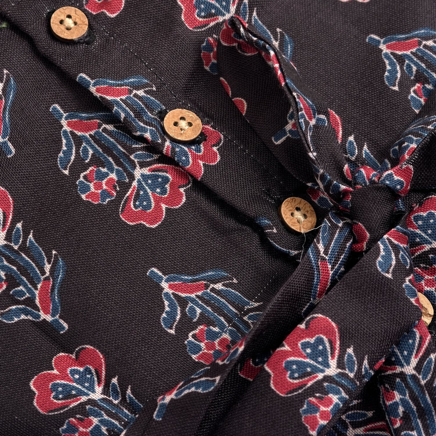 Black sleeveless slub cotton frock for baby girls with red floral block prints and a pleated flare, displayed flat on a white fur background with floral basket hanger.