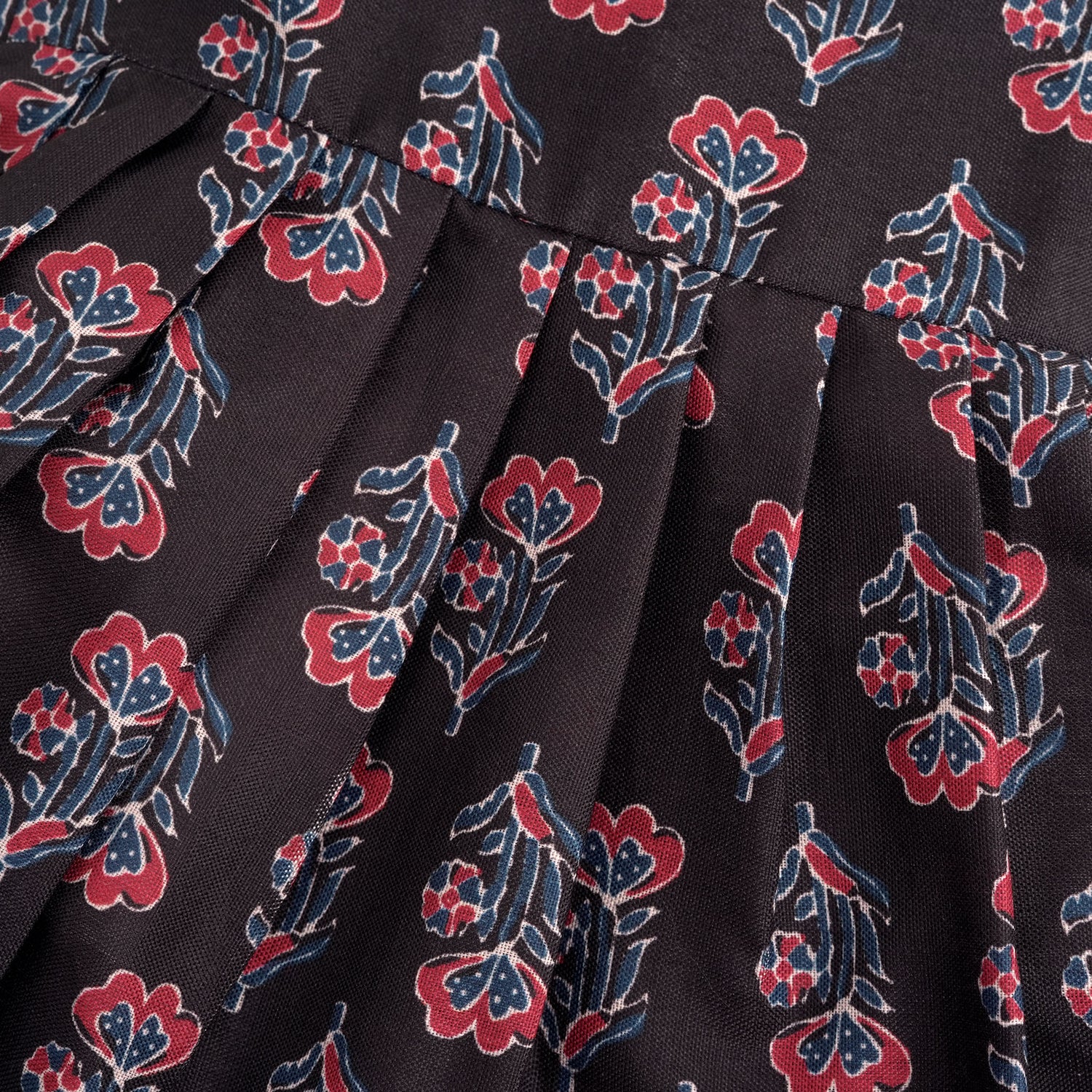 Black sleeveless slub cotton frock for baby girls with red floral block prints and a pleated flare, displayed flat on a white fur background with floral basket hanger.