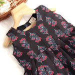 Black sleeveless slub cotton frock for baby girls with red floral block prints and a pleated flare, displayed flat on a white fur background with floral basket hanger.