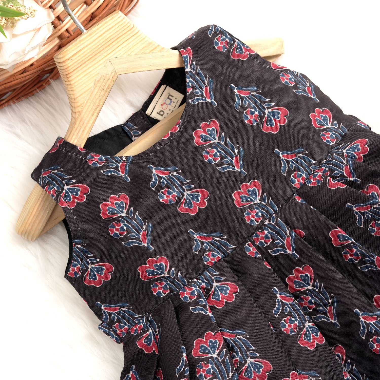 Black sleeveless slub cotton frock for baby girls with red floral block prints and a pleated flare, displayed flat on a white fur background with floral basket hanger.