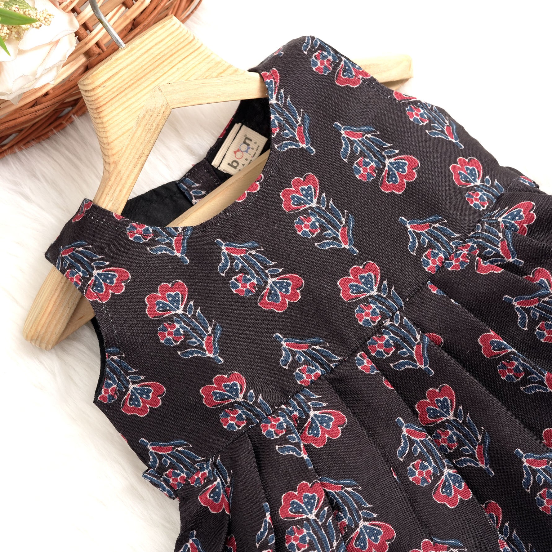 Black sleeveless slub cotton frock for baby girls with red floral block prints and a pleated flare, displayed flat on a white fur background with floral basket hanger.