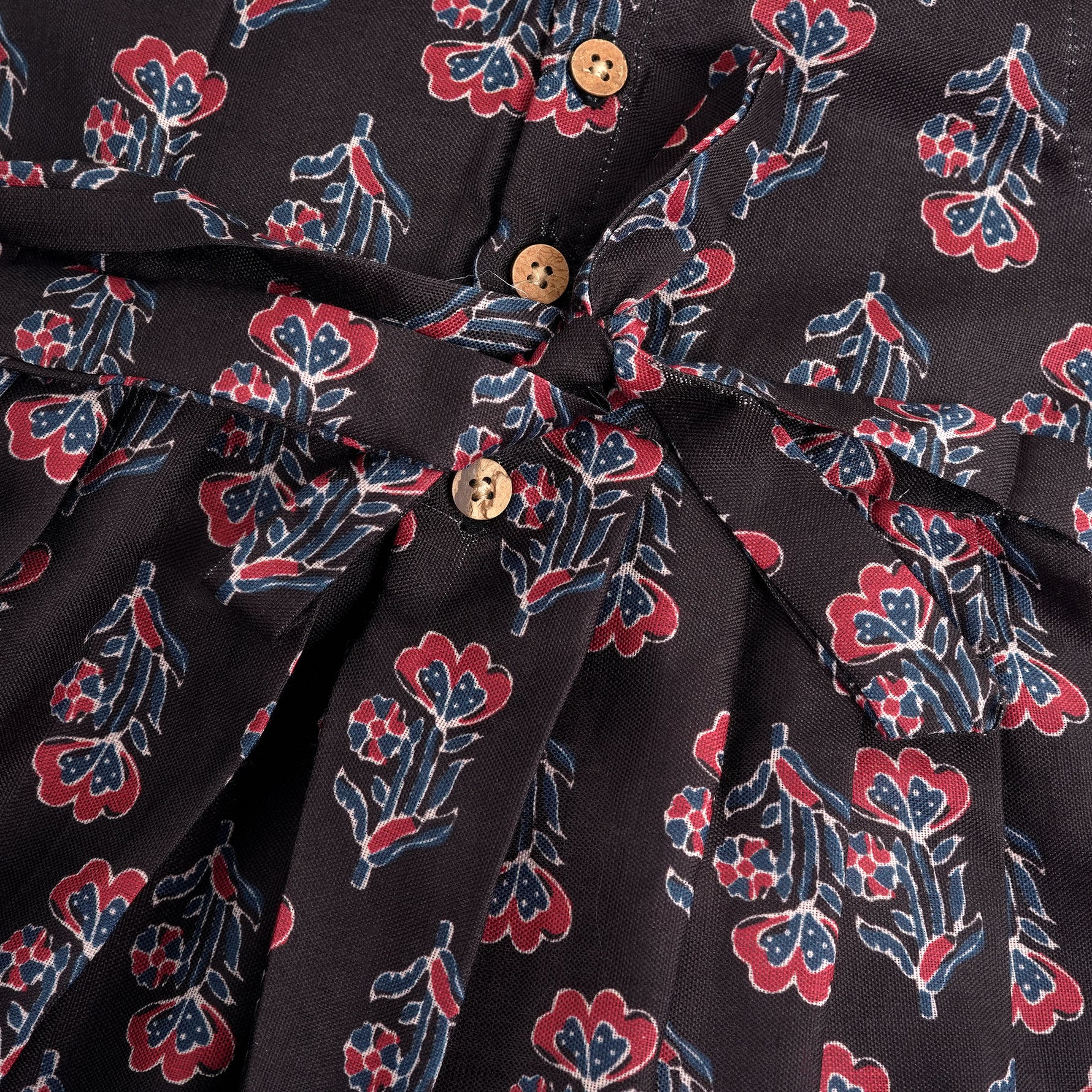 Black sleeveless slub cotton frock for baby girls with red floral block prints and a pleated flare, displayed flat on a white fur background with floral basket hanger.