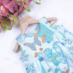 Girls' sleeveless frock in pastel blue cotton with butterfly and floral print, displayed with a wooden hanger under pink orchids – Boon Babies summer casual wear collection.
