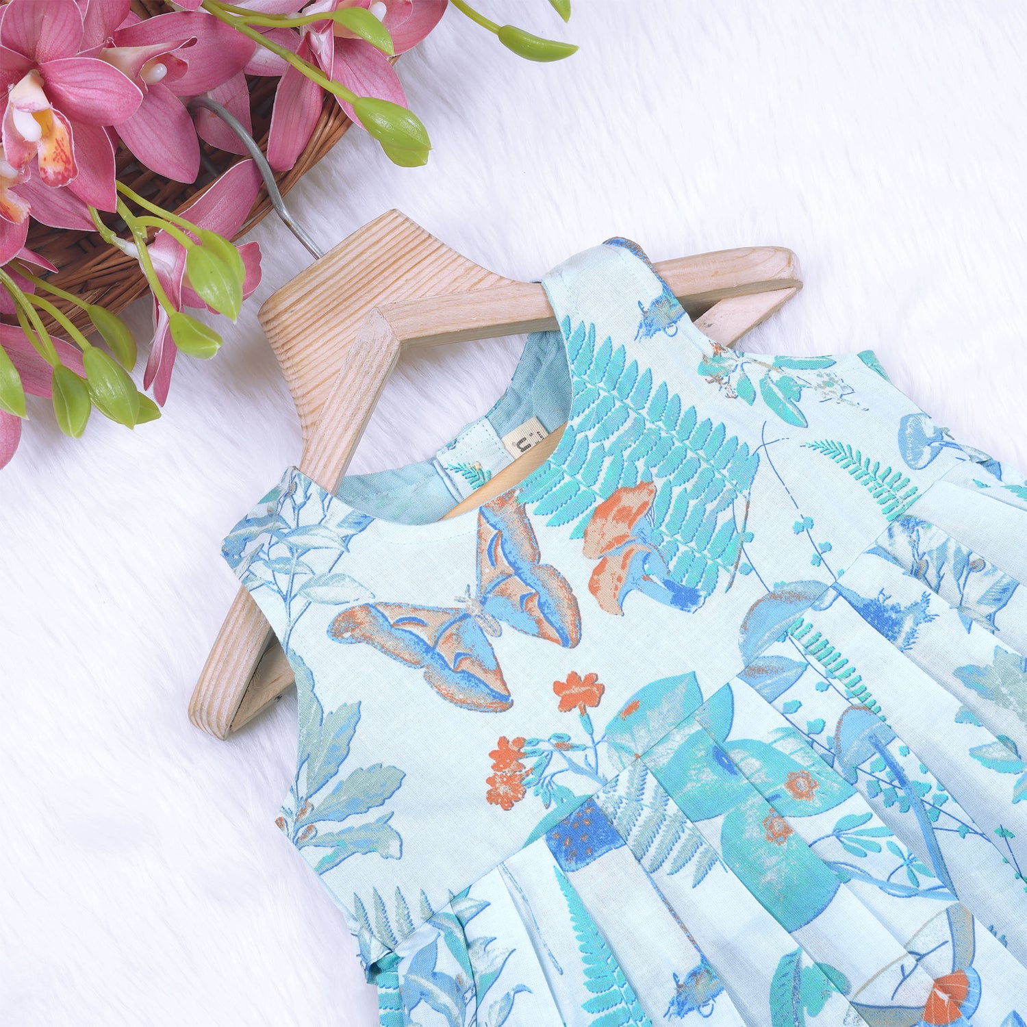 Girls' sleeveless frock in pastel blue cotton with butterfly and floral print, displayed with a wooden hanger under pink orchids – Boon Babies summer casual wear collection.
