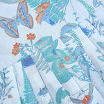 Girls' sleeveless frock in pastel blue cotton with butterfly and floral print, displayed with a wooden hanger under pink orchids – Boon Babies summer casual wear collection.
