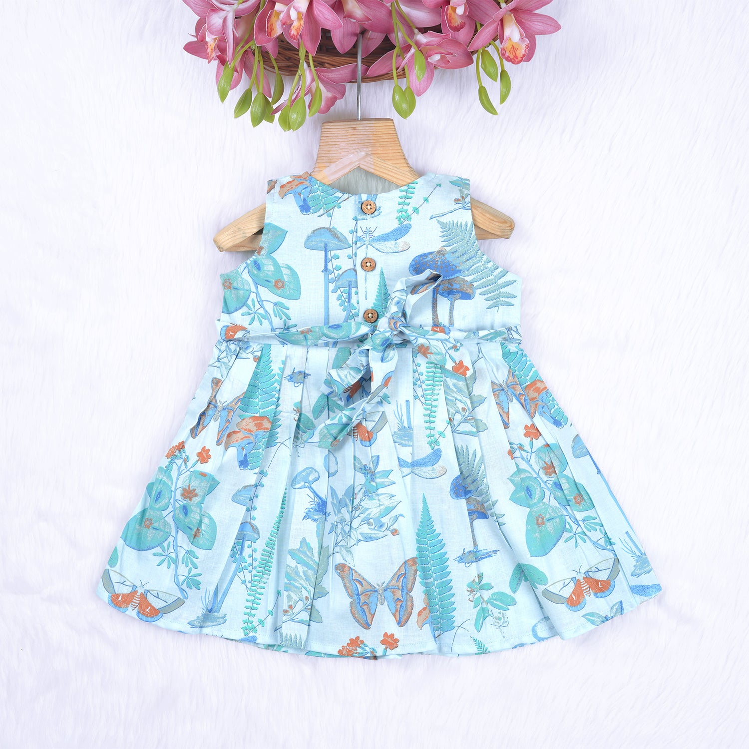 Girls' sleeveless frock in pastel blue cotton with butterfly and floral print, displayed with a wooden hanger under pink orchids – Boon Babies summer casual wear collection.

