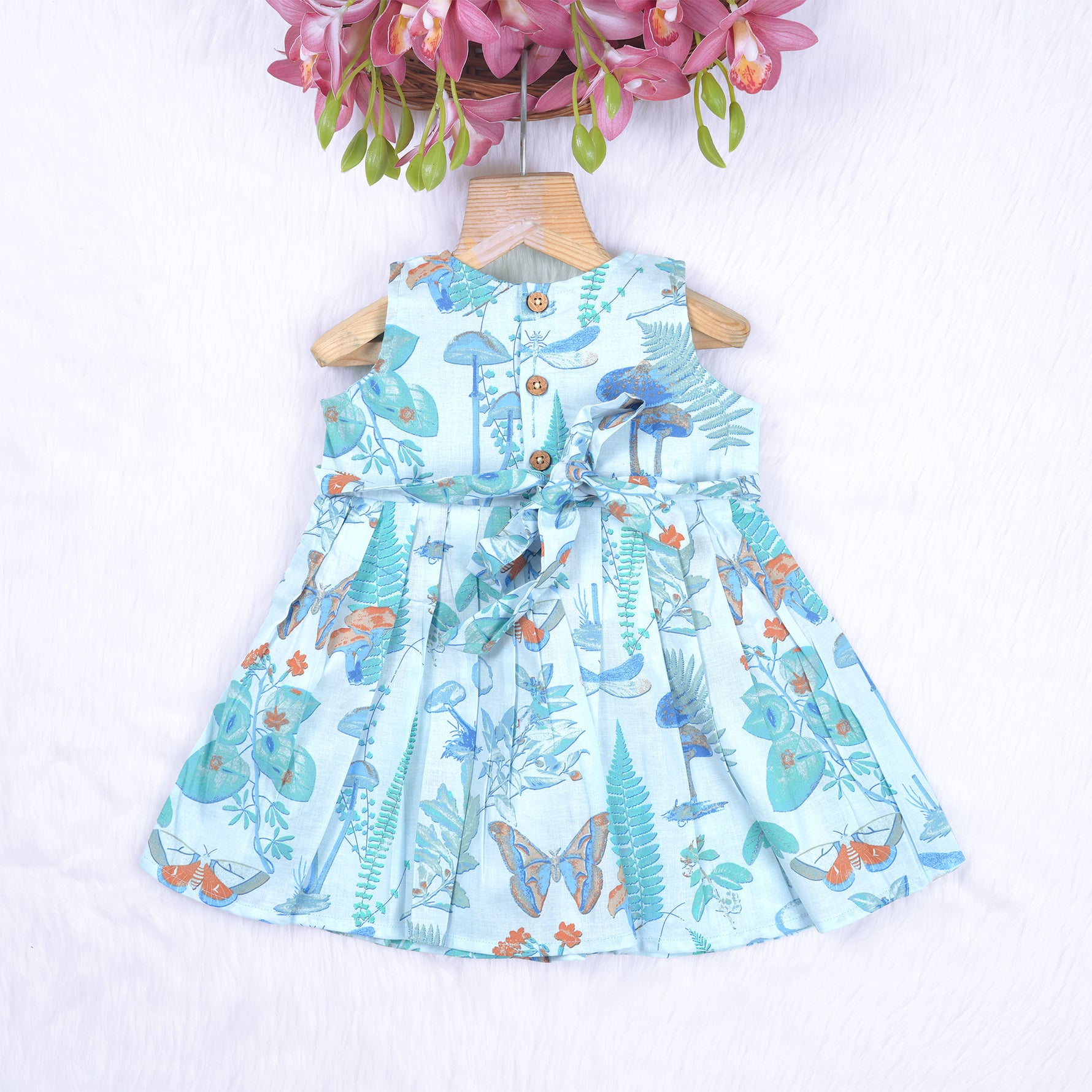Girls' sleeveless frock in pastel blue cotton with butterfly and floral print, displayed with a wooden hanger under pink orchids – Boon Babies summer casual wear collection.
