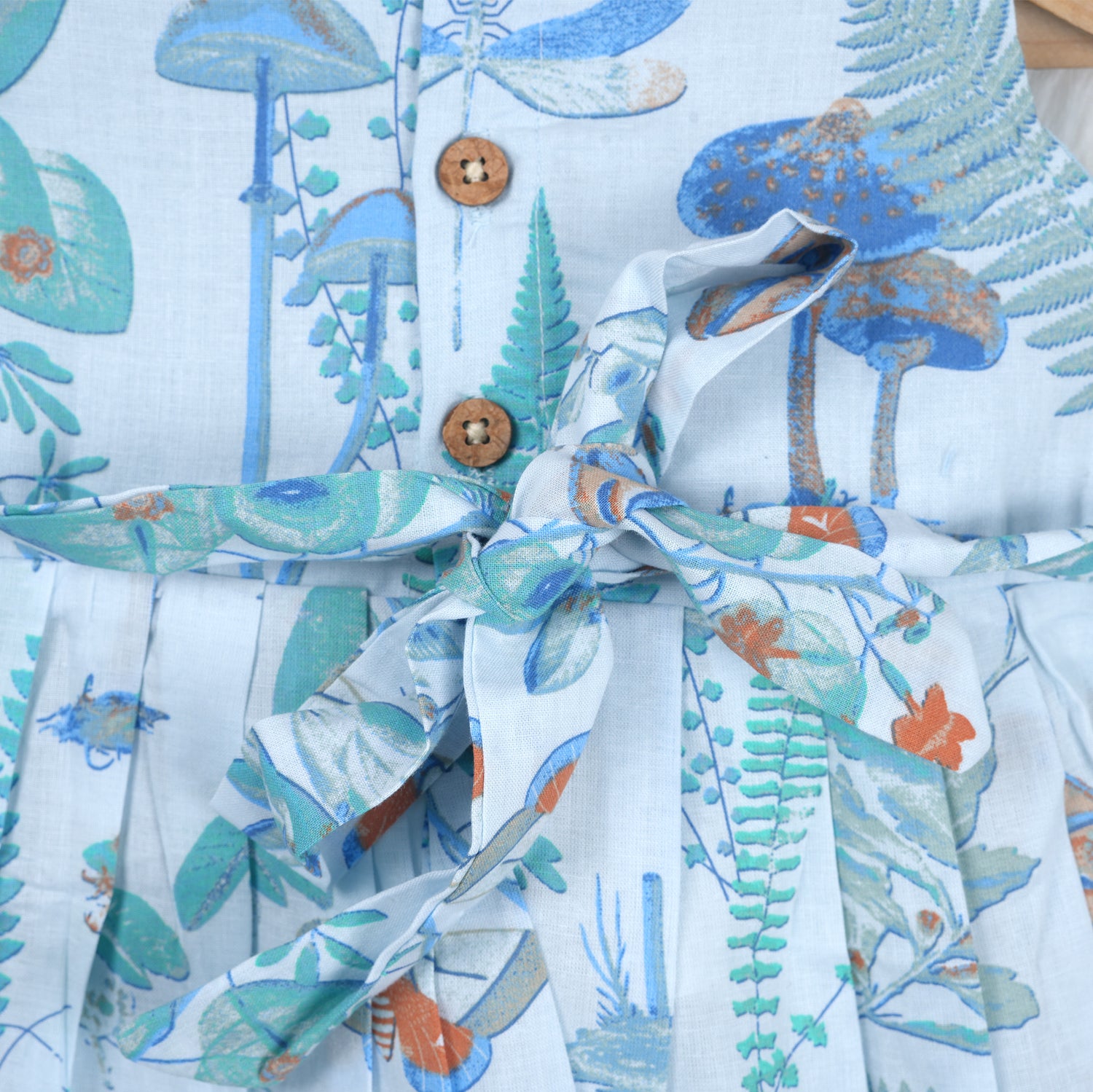 Girls' sleeveless frock in pastel blue cotton with butterfly and floral print, displayed with a wooden hanger under pink orchids – Boon Babies summer casual wear collection.
