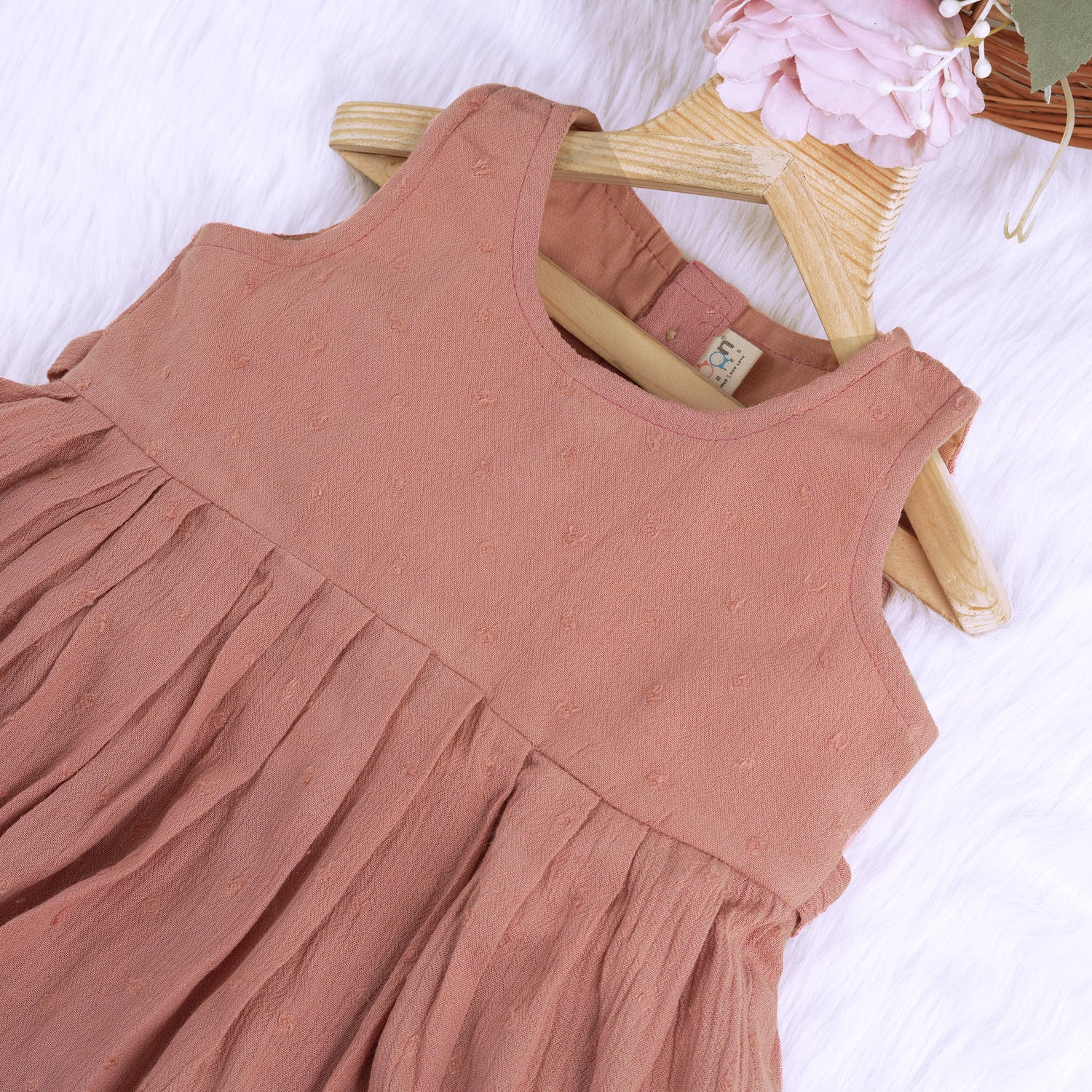 Brown crinkled cotton sleeveless frock for baby girls with pleated skirt design, displayed on a wooden hanger over a white faux fur background with floral accents.