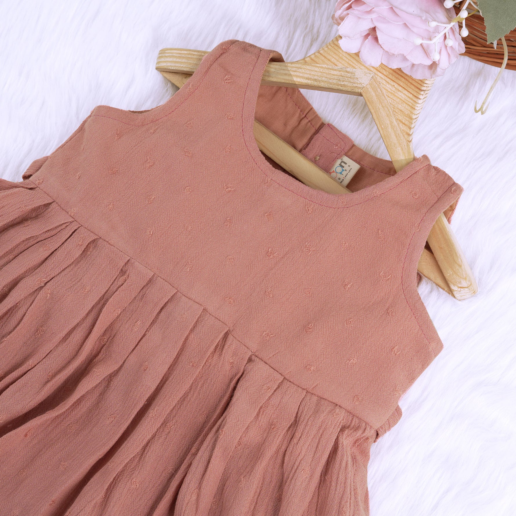 Brown crinkled cotton sleeveless frock for baby girls with pleated skirt design, displayed on a wooden hanger over a white faux fur background with floral accents.