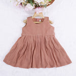 Brown crinkled cotton sleeveless frock for baby girls with pleated skirt design, displayed on a wooden hanger over a white faux fur background with floral accents.