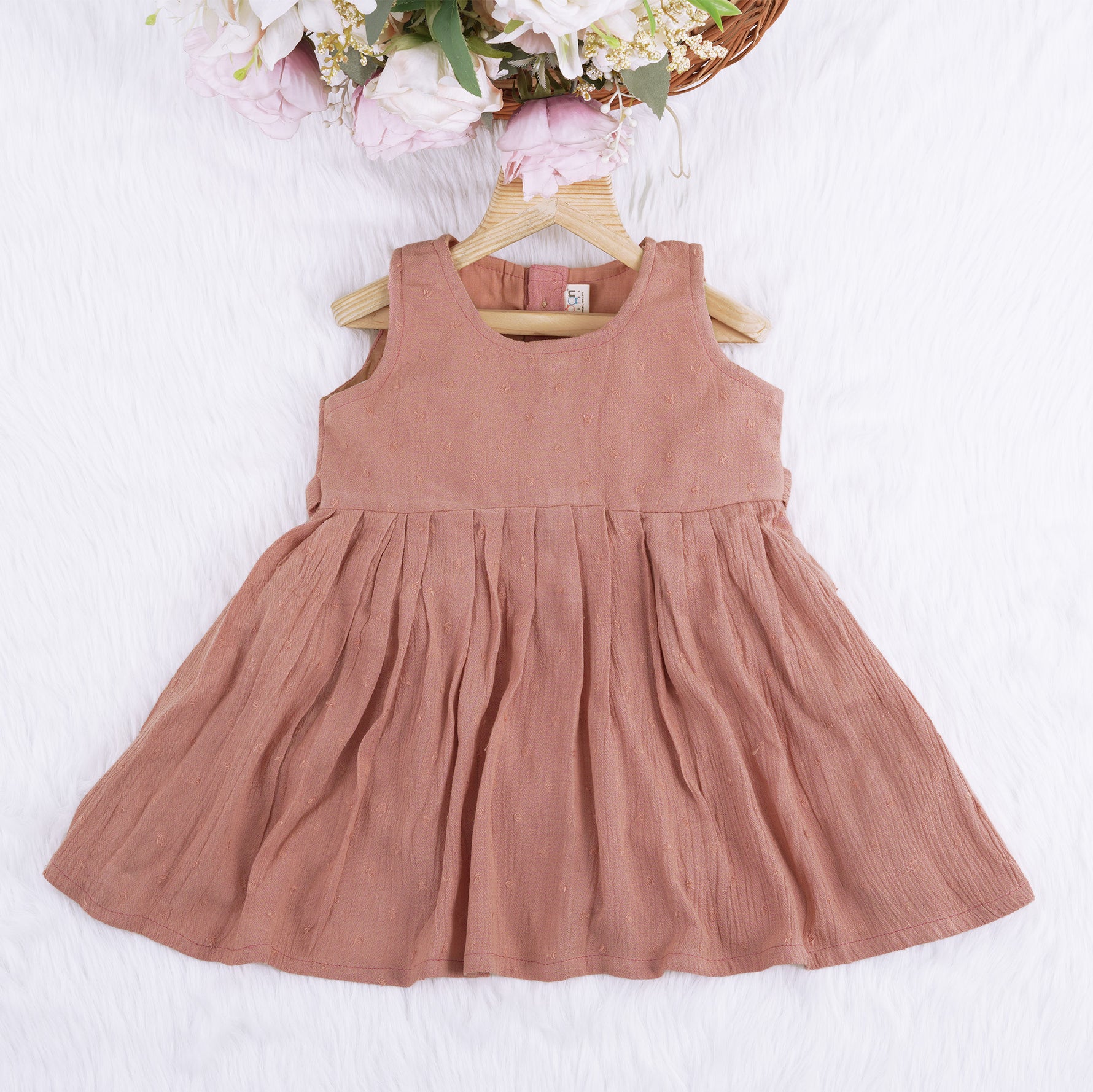 Brown crinkled cotton sleeveless frock for baby girls with pleated skirt design, displayed on a wooden hanger over a white faux fur background with floral accents.