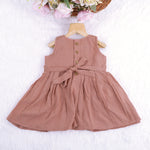 Brown crinkled cotton sleeveless frock for baby girls with pleated skirt design, displayed on a wooden hanger over a white faux fur background with floral accents.