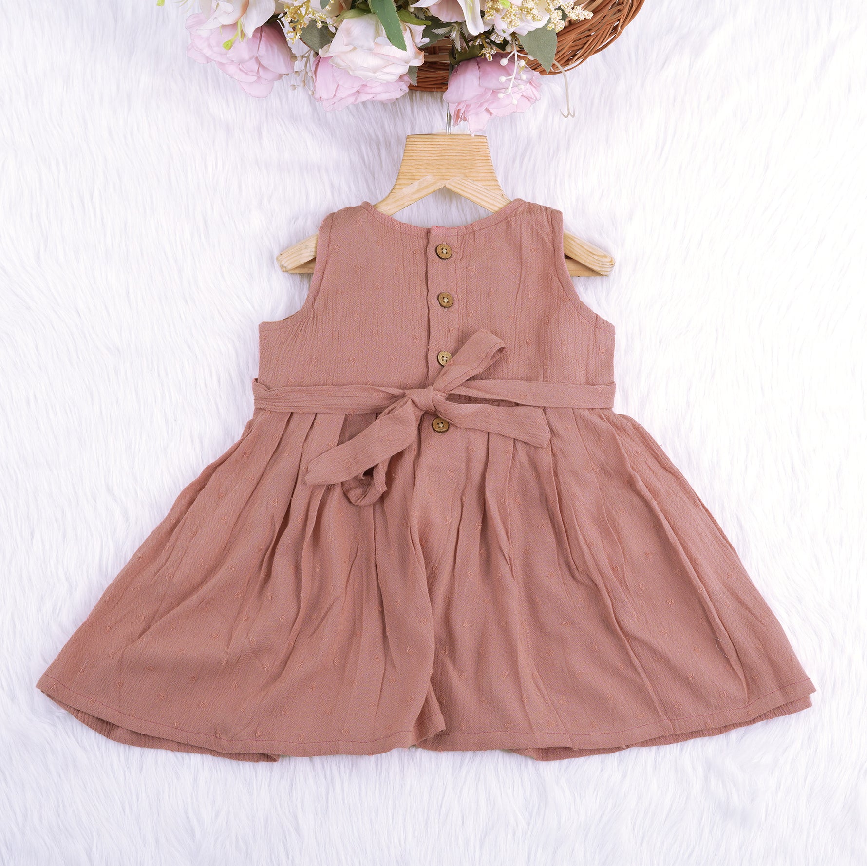 Brown crinkled cotton sleeveless frock for baby girls with pleated skirt design, displayed on a wooden hanger over a white faux fur background with floral accents.