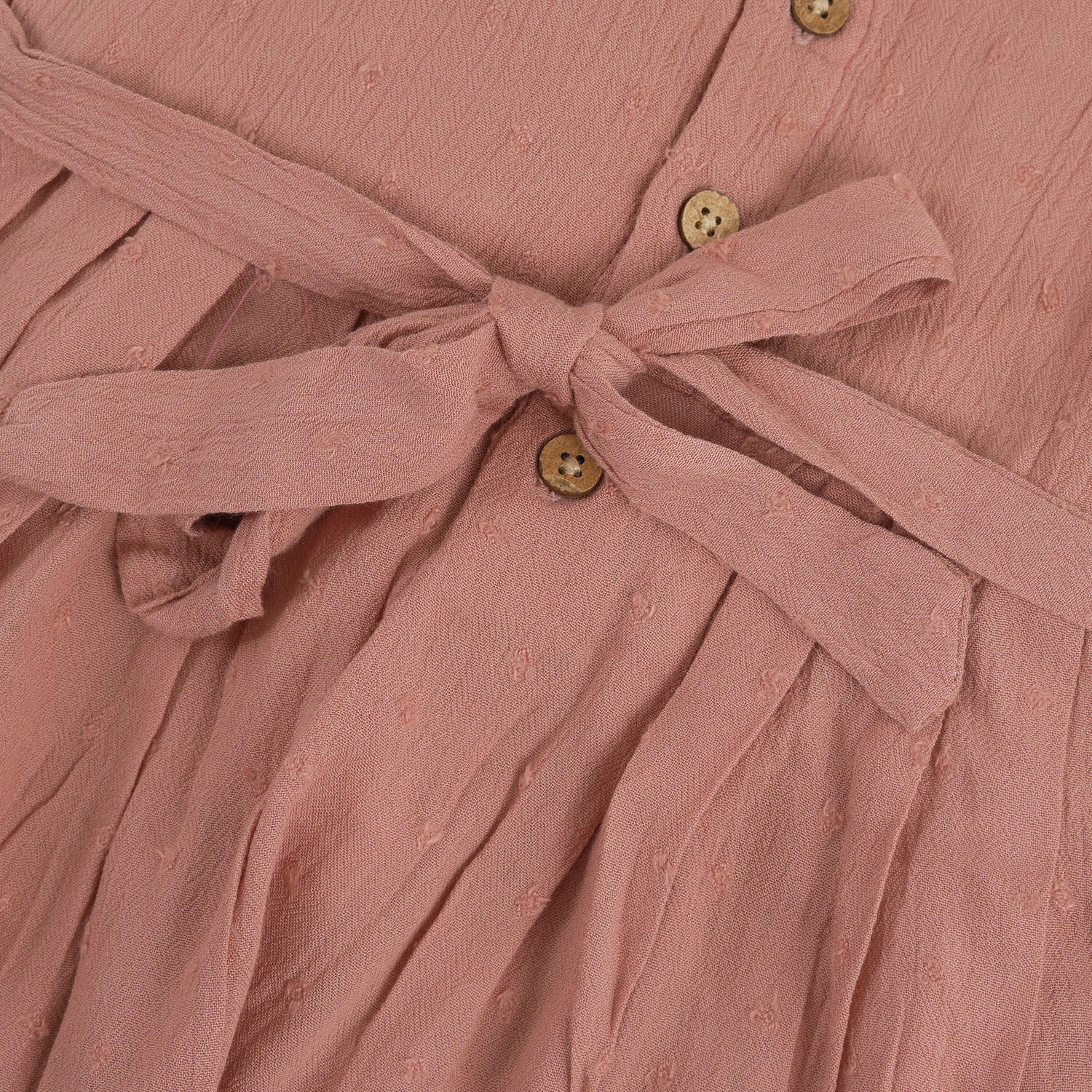 Brown crinkled cotton sleeveless frock for baby girls with pleated skirt design, displayed on a wooden hanger over a white faux fur background with floral accents.