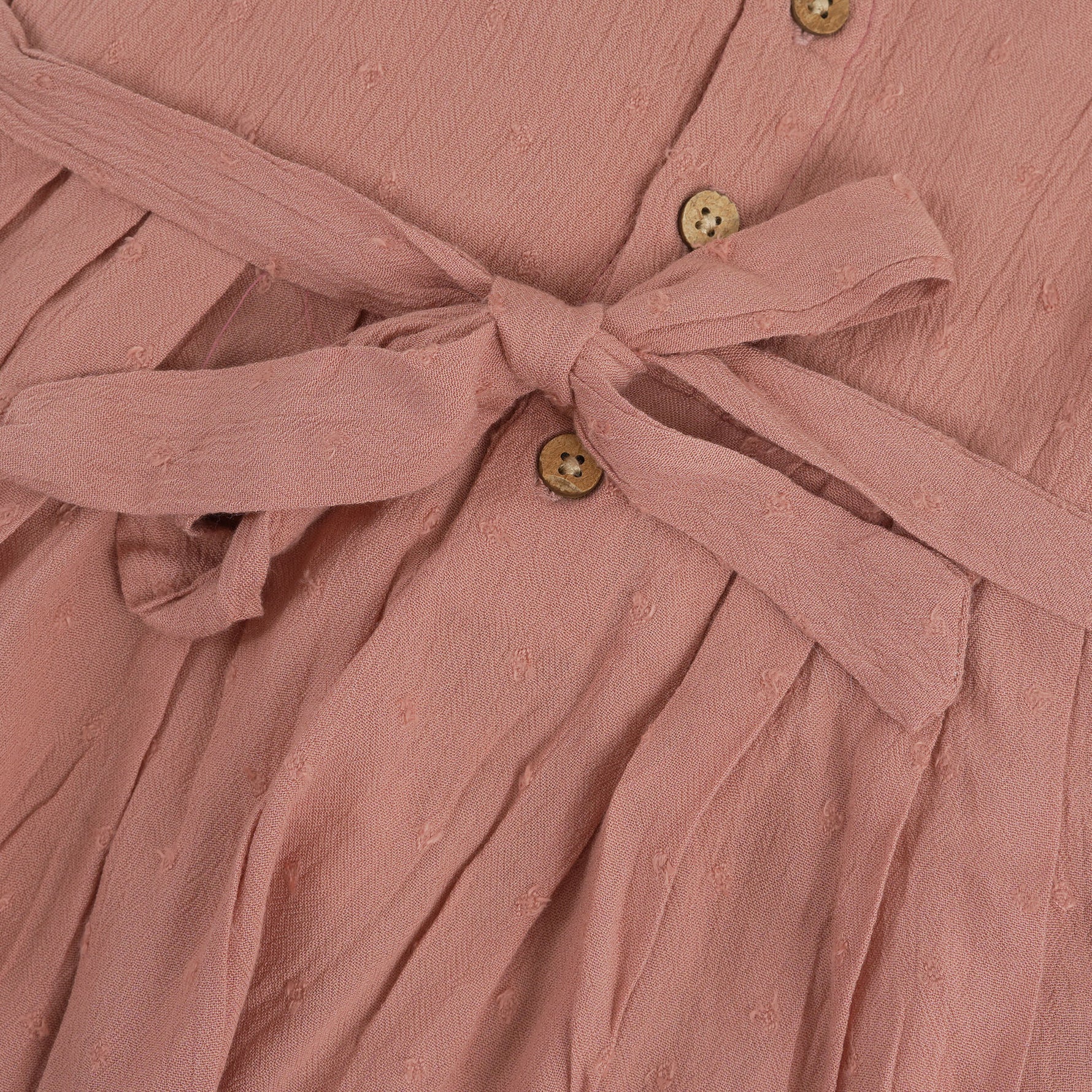 Brown crinkled cotton sleeveless frock for baby girls with pleated skirt design, displayed on a wooden hanger over a white faux fur background with floral accents.
