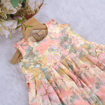 Floral printed sleeveless frock for baby girls in pastel shades of pink, beige, and grey, made from chiffon cotton and displayed on a wooden hanger over white fur with flower props.