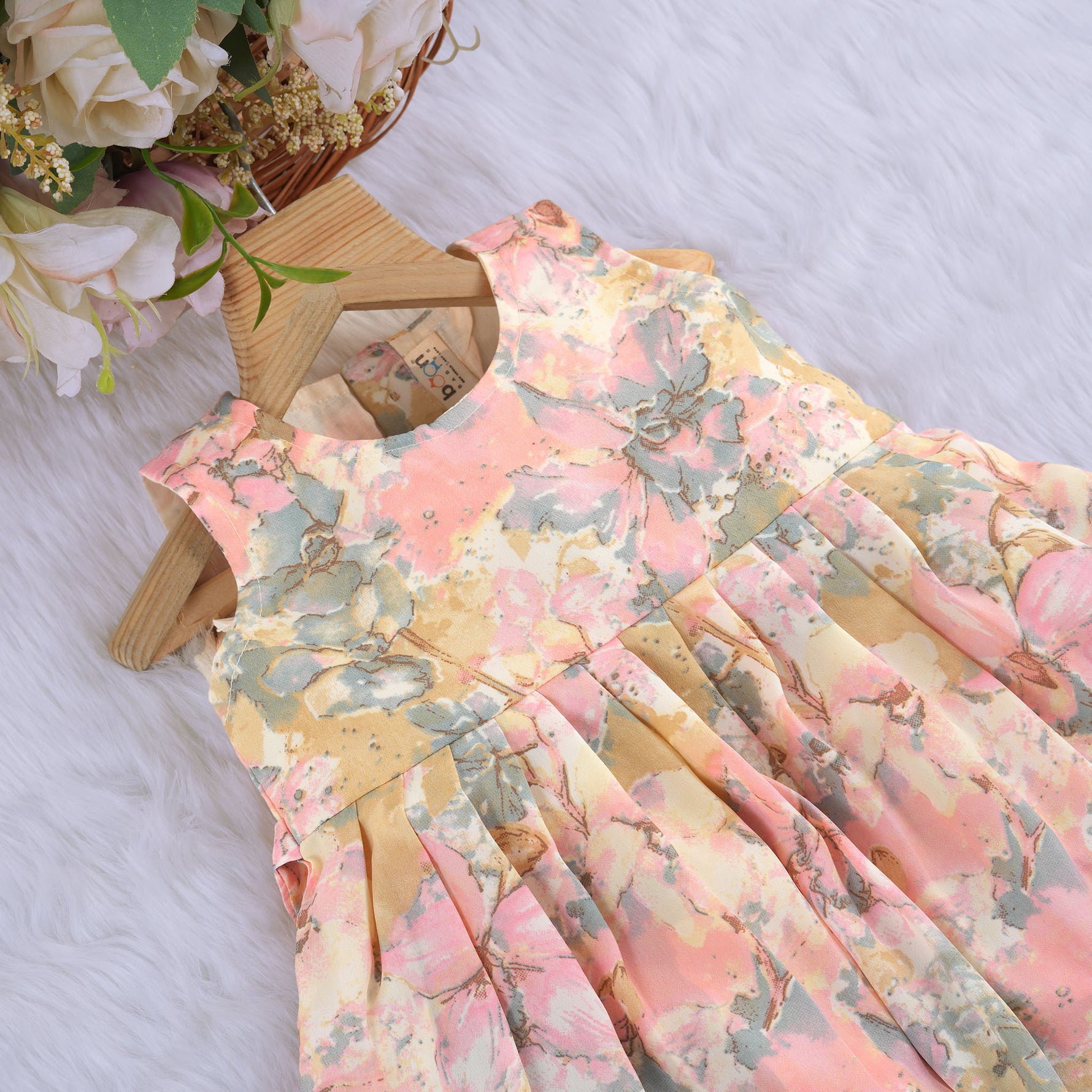 Floral printed sleeveless frock for baby girls in pastel shades of pink, beige, and grey, made from chiffon cotton and displayed on a wooden hanger over white fur with flower props.