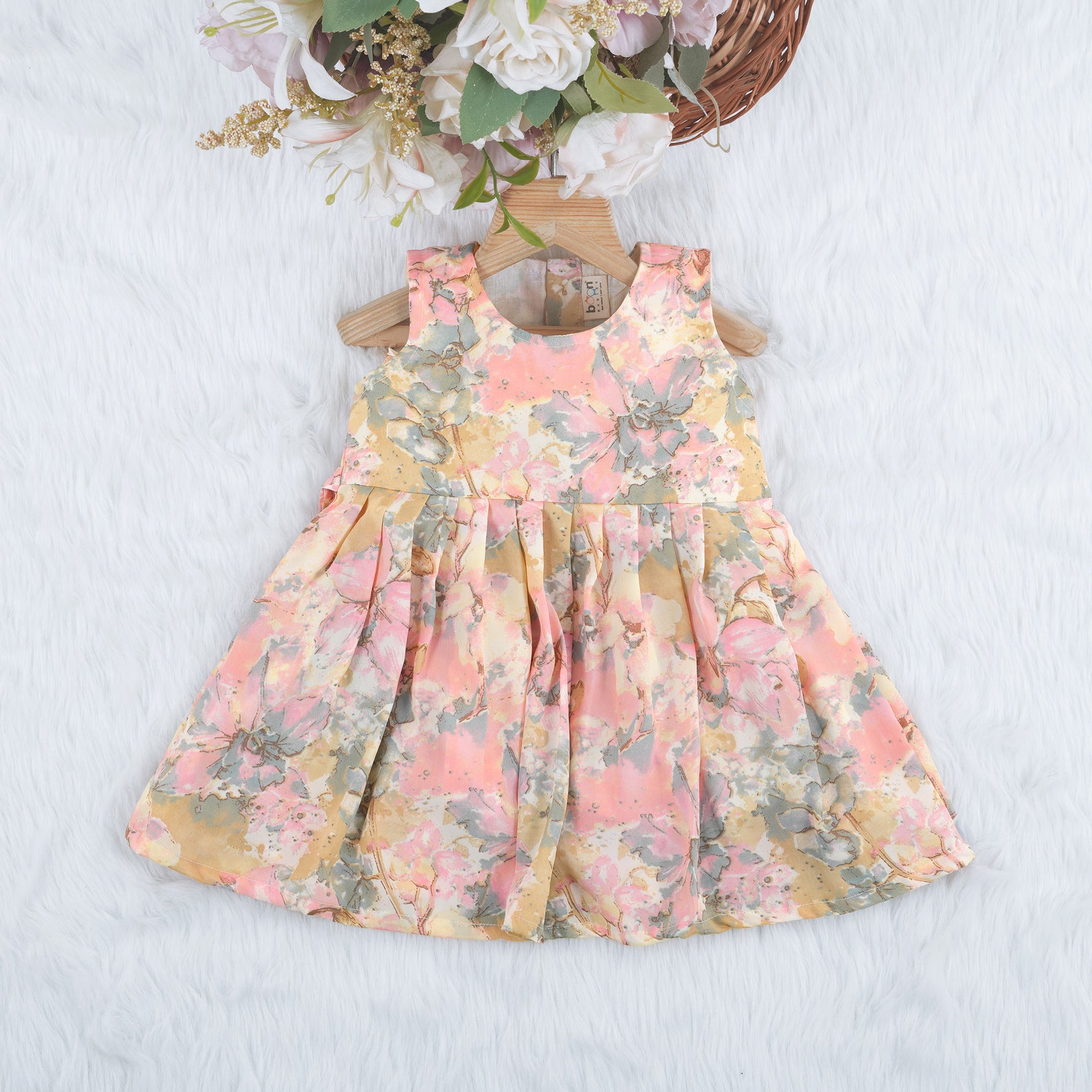 Floral printed sleeveless frock for baby girls in pastel shades of pink, beige, and grey, made from chiffon cotton and displayed on a wooden hanger over white fur with flower props.