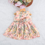 Floral printed sleeveless frock for baby girls in pastel shades of pink, beige, and grey, made from chiffon cotton and displayed on a wooden hanger over white fur with flower props.