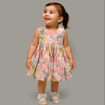 Floral printed sleeveless frock for baby girls in pastel shades of pink, beige, and grey, made from chiffon cotton and displayed on a wooden hanger over white fur with flower props.