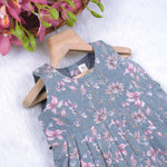 Sleeveless grey cotton frock with pink floral prints, neatly pleated and displayed on a wooden hanger under fresh orchids – from Boon Babies girlswear collection.
