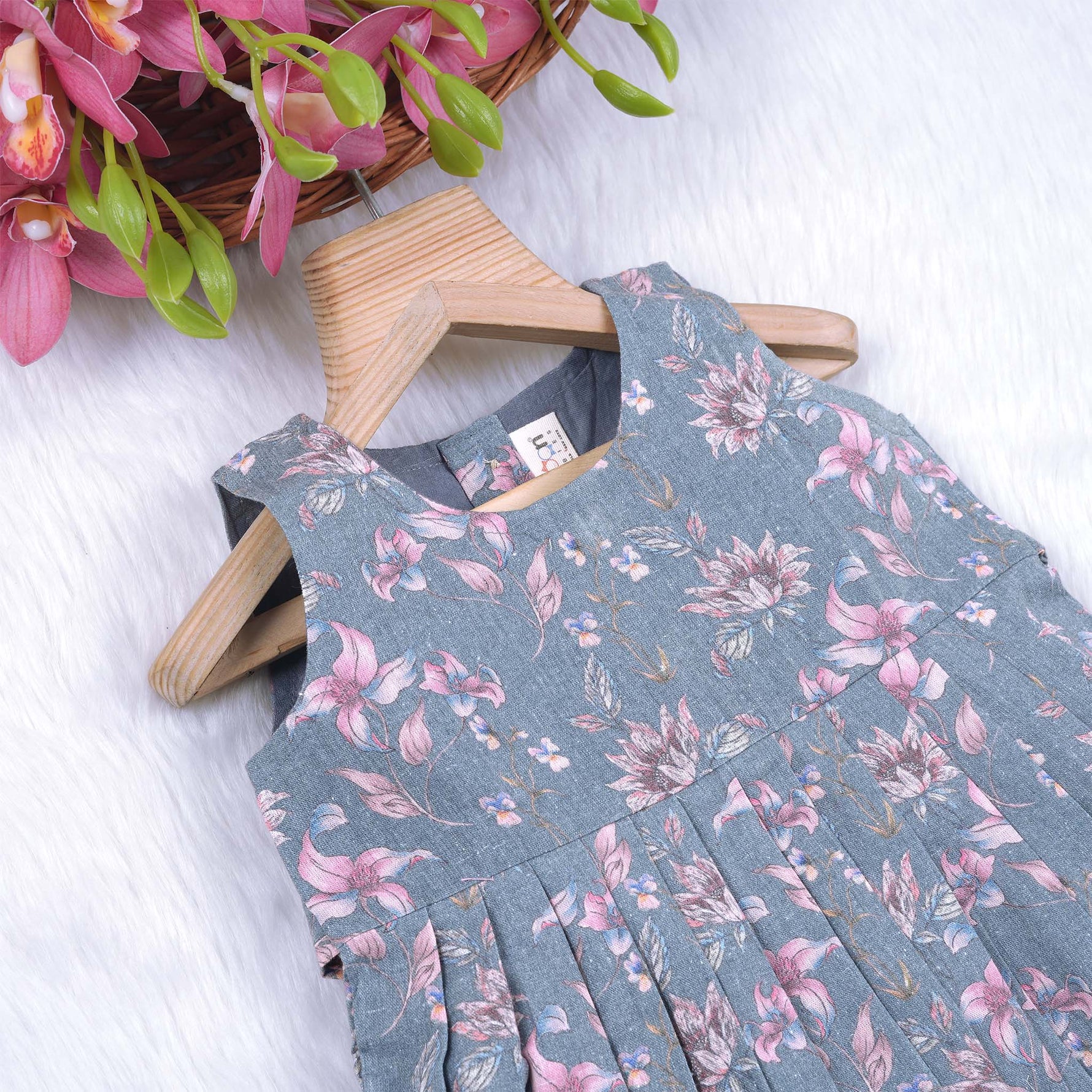 Sleeveless grey cotton frock with pink floral prints, neatly pleated and displayed on a wooden hanger under fresh orchids – from Boon Babies girlswear collection.

