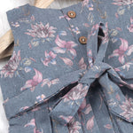 Sleeveless grey cotton frock with pink floral prints, neatly pleated and displayed on a wooden hanger under fresh orchids – from Boon Babies girlswear collection.
