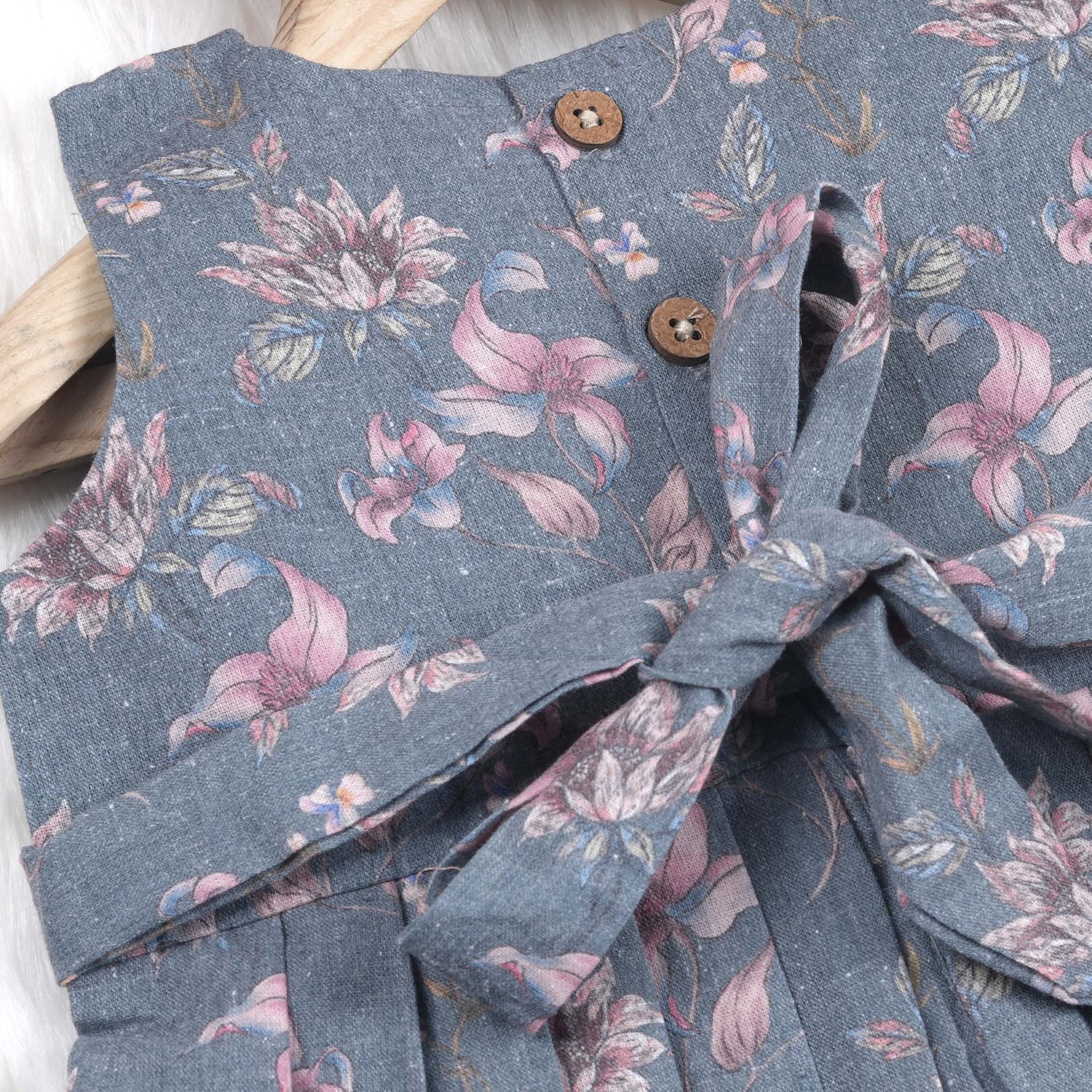 Sleeveless grey cotton frock with pink floral prints, neatly pleated and displayed on a wooden hanger under fresh orchids – from Boon Babies girlswear collection.
