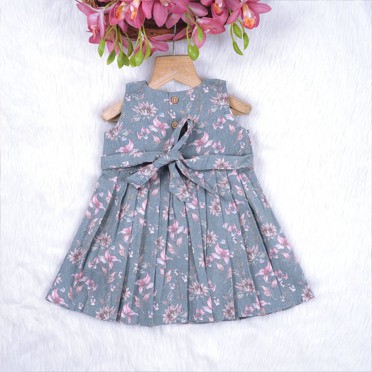 Sleeveless grey cotton frock with pink floral prints, neatly pleated and displayed on a wooden hanger under fresh orchids – from Boon Babies girlswear collection.
