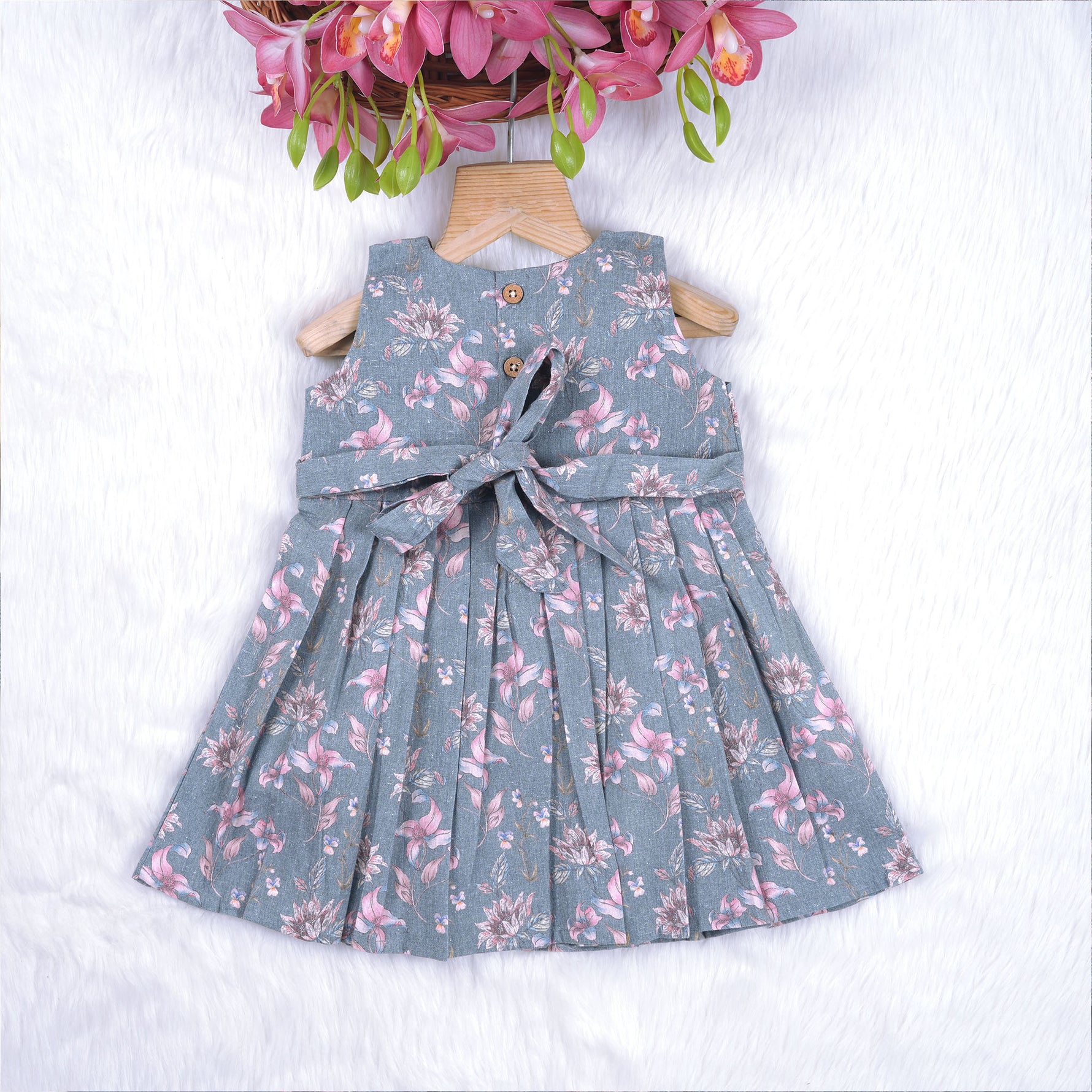 Sleeveless grey cotton frock with pink floral prints, neatly pleated and displayed on a wooden hanger under fresh orchids – from Boon Babies girlswear collection.
