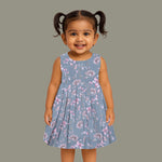Sleeveless grey cotton frock with pink floral prints, neatly pleated and displayed on a wooden hanger under fresh orchids – from Boon Babies girlswear collection.
