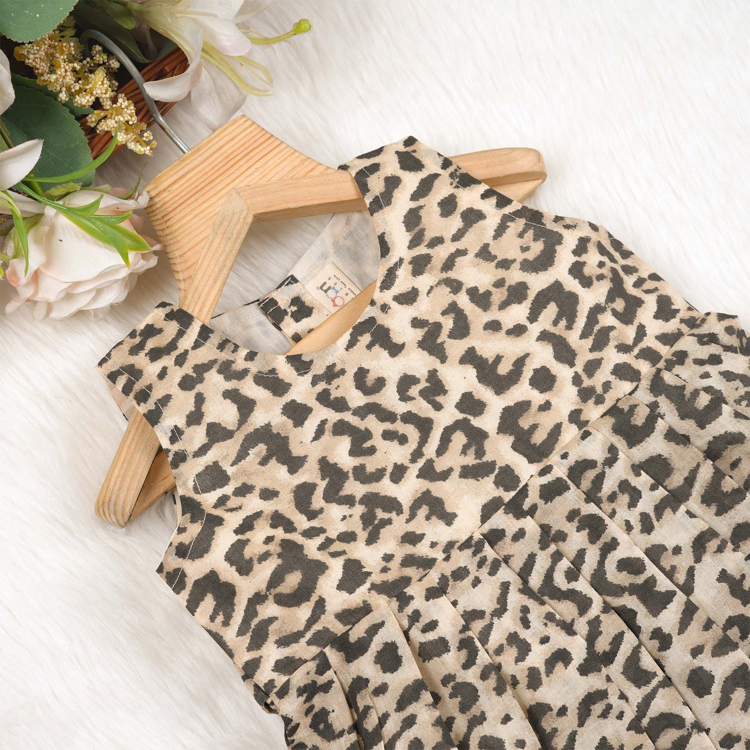 Leopard print sleeveless cotton frock for girls aged 12M–5Y, with a flared pleated skirt – displayed on a wooden hanger with white roses in the background.