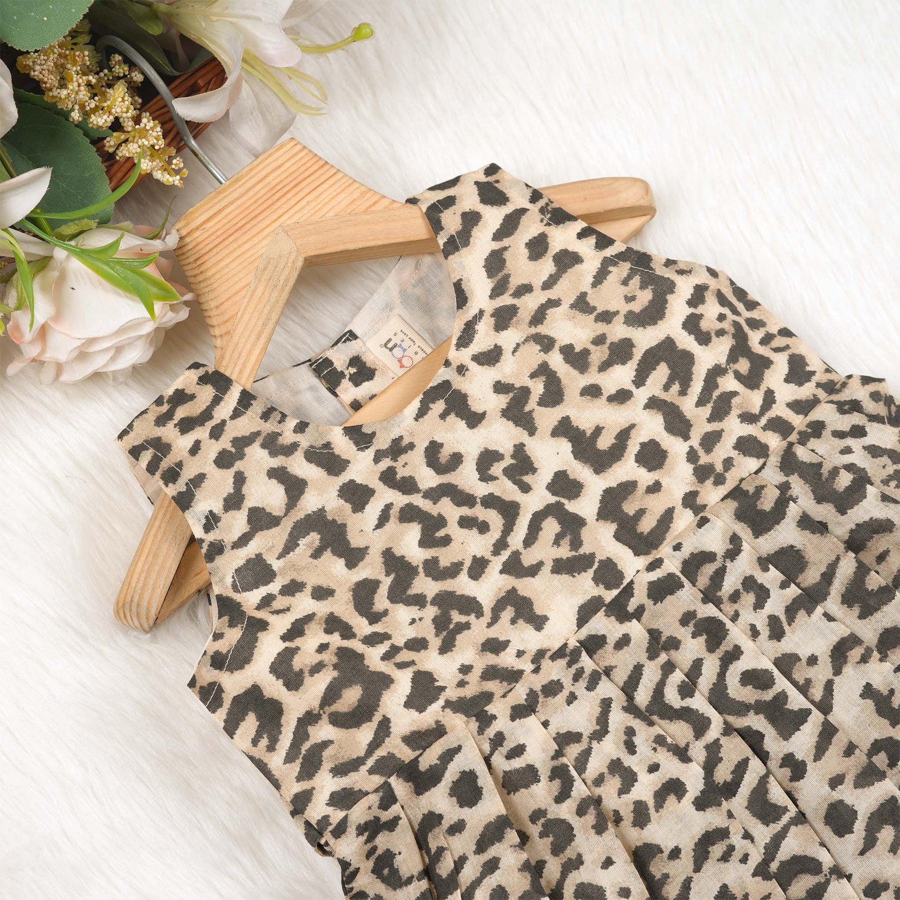 Leopard print sleeveless cotton frock for girls aged 12M–5Y, with a flared pleated skirt – displayed on a wooden hanger with white roses in the background.